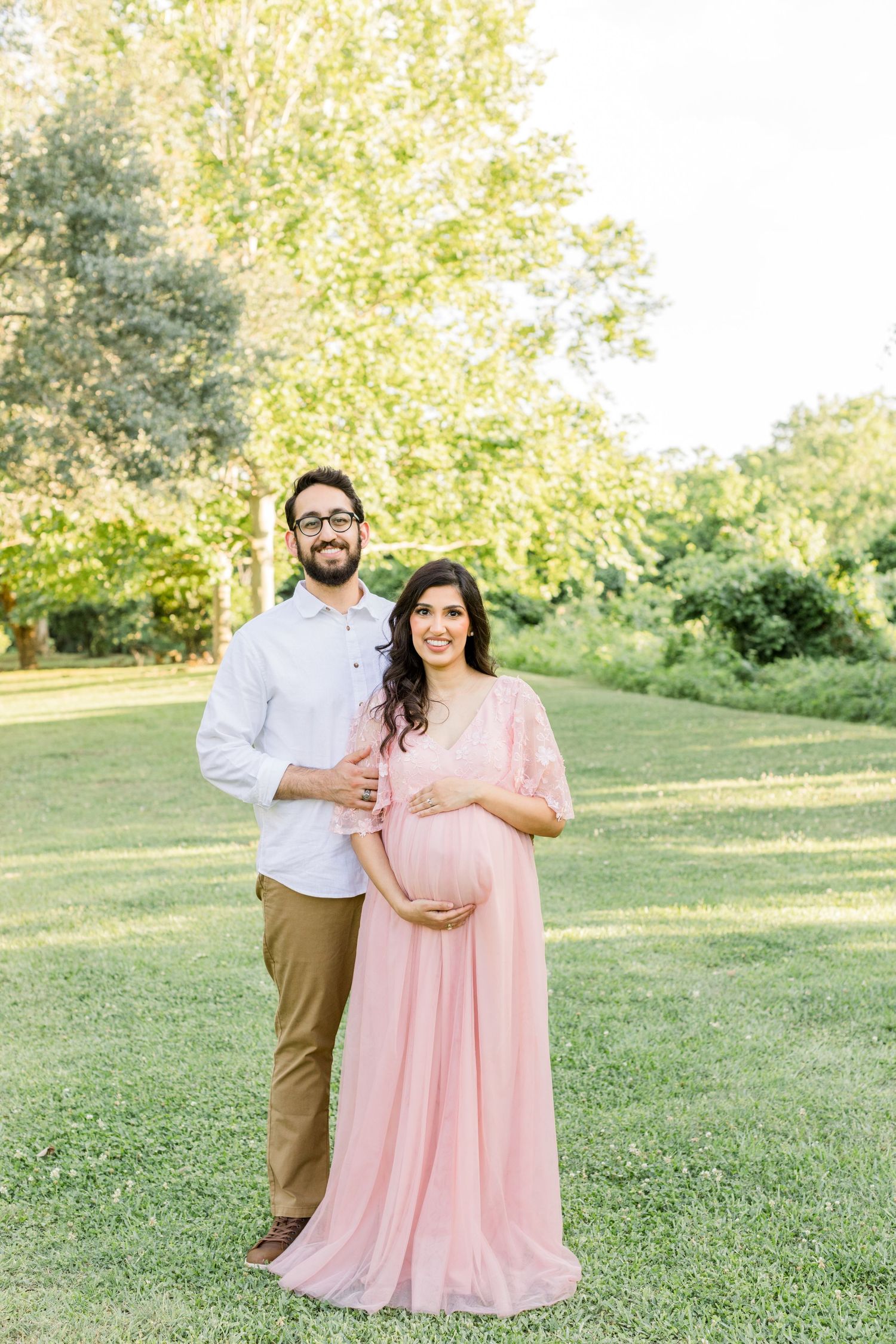 Couple poses in sunlit garden setting with woman wearing flowing pink maternity dress.