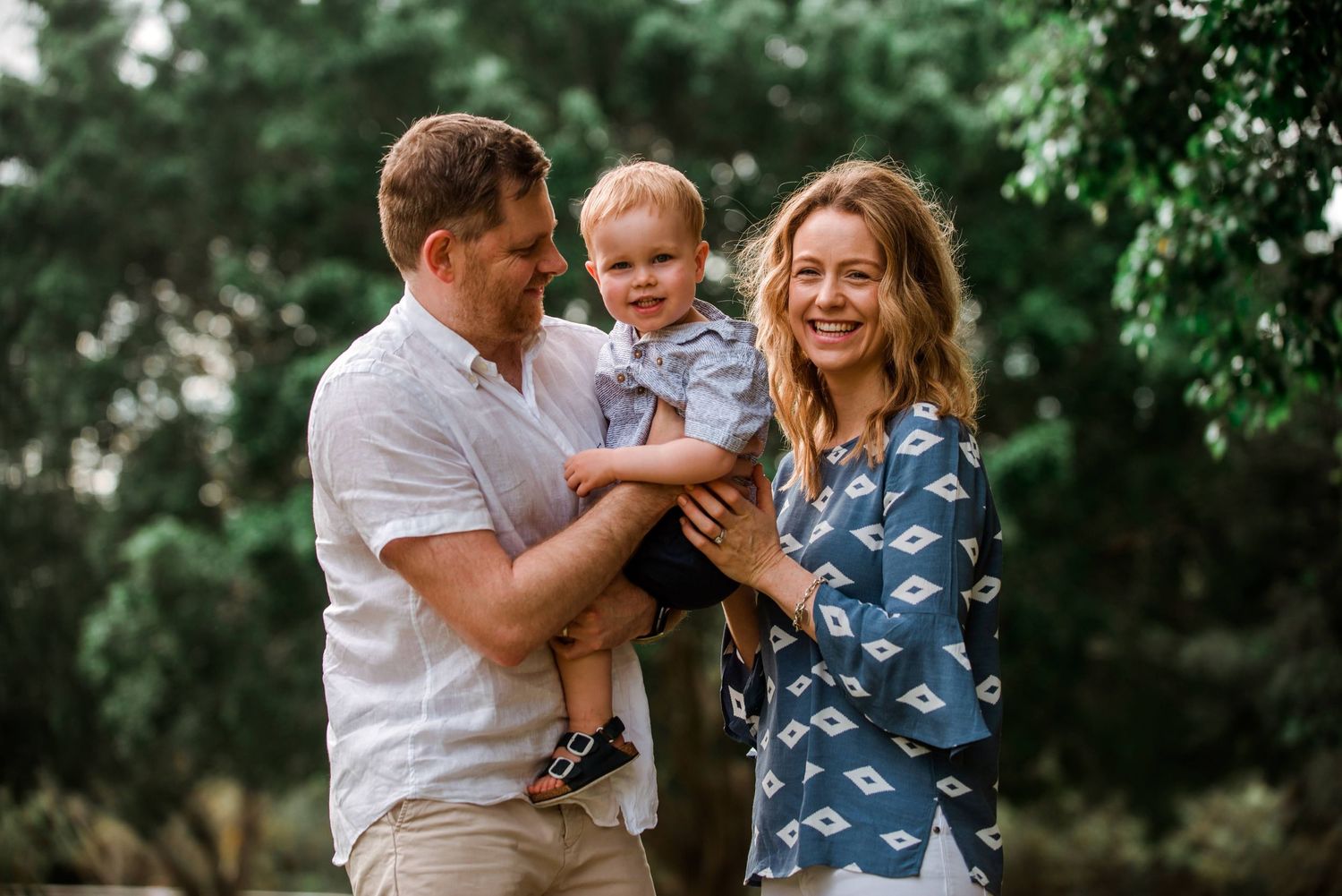 Family of three shares a happy moment together outdoors against green foliage.