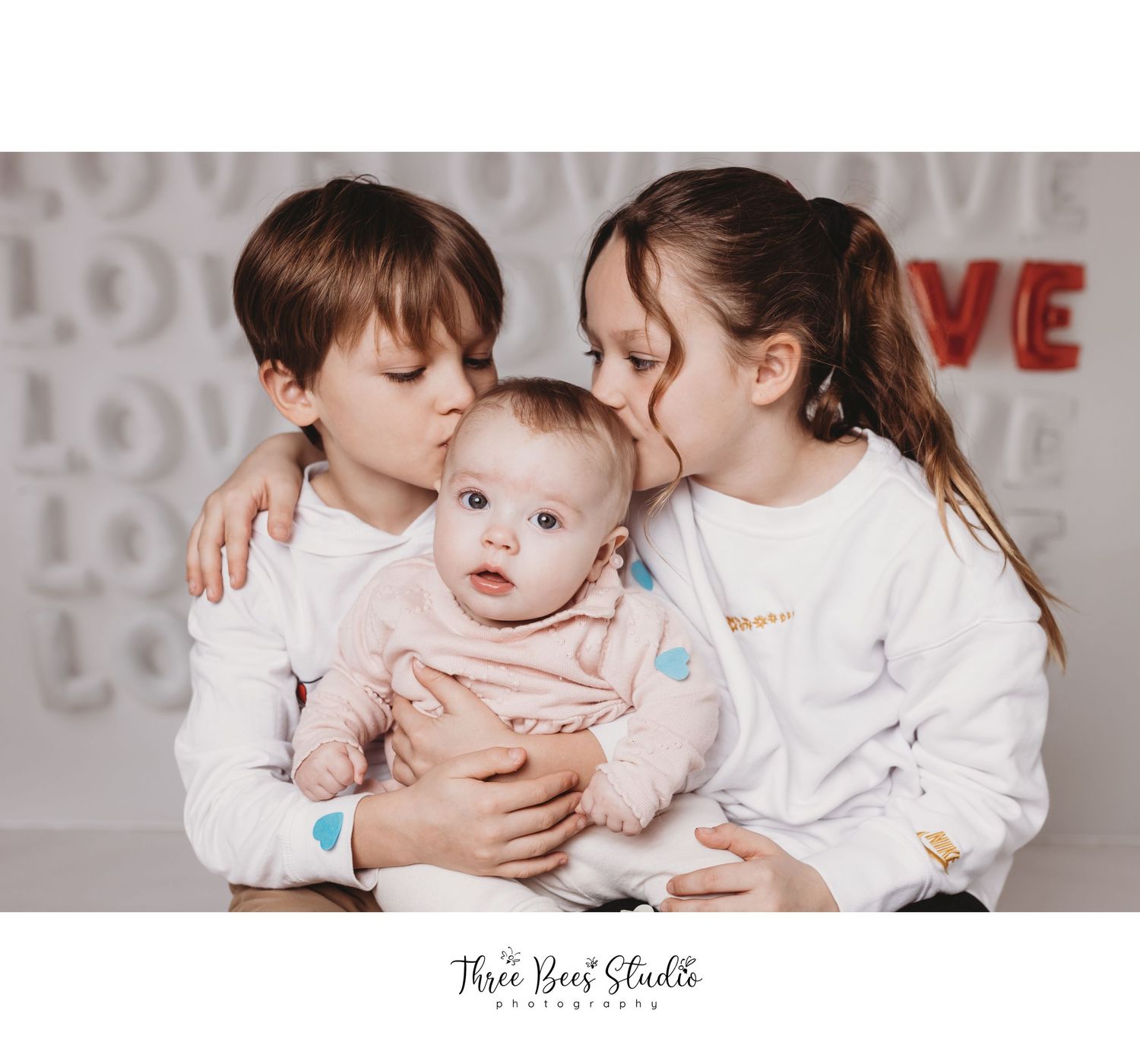 Family portrait with siblings and baby against love-themed backdrop.