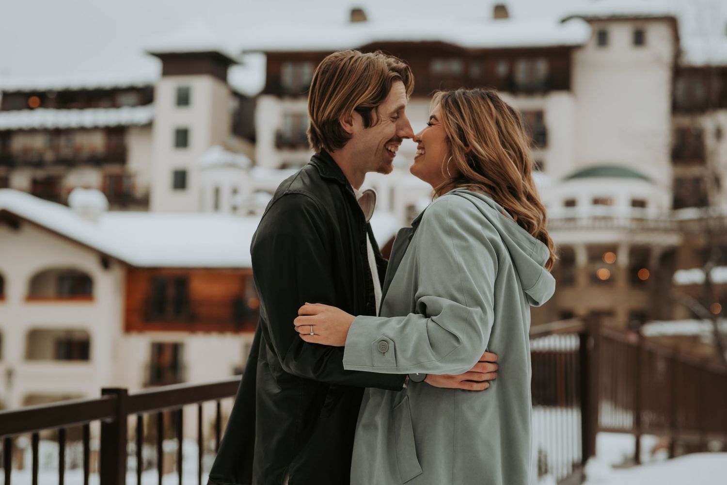 Smiling couple embraces and touch noses on a snowy balcony overlooking a winter mountain village lodge.