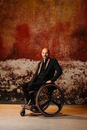 Professional in a business suit sits confidently in a wheelchair against a rustic red wall.