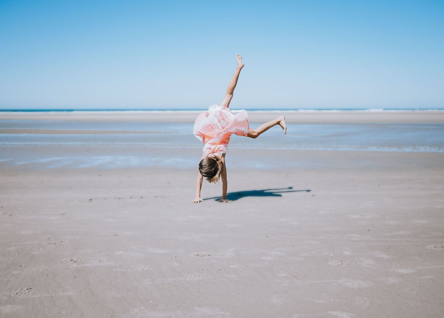 Person performing a cartwheel in pink dress against blue ocean backdrop.