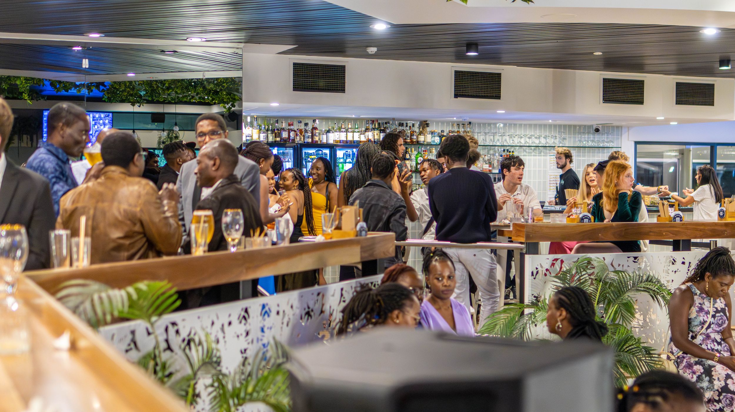 Crowded modern bar or restaurant interior with people gathering around tables and counter seating.