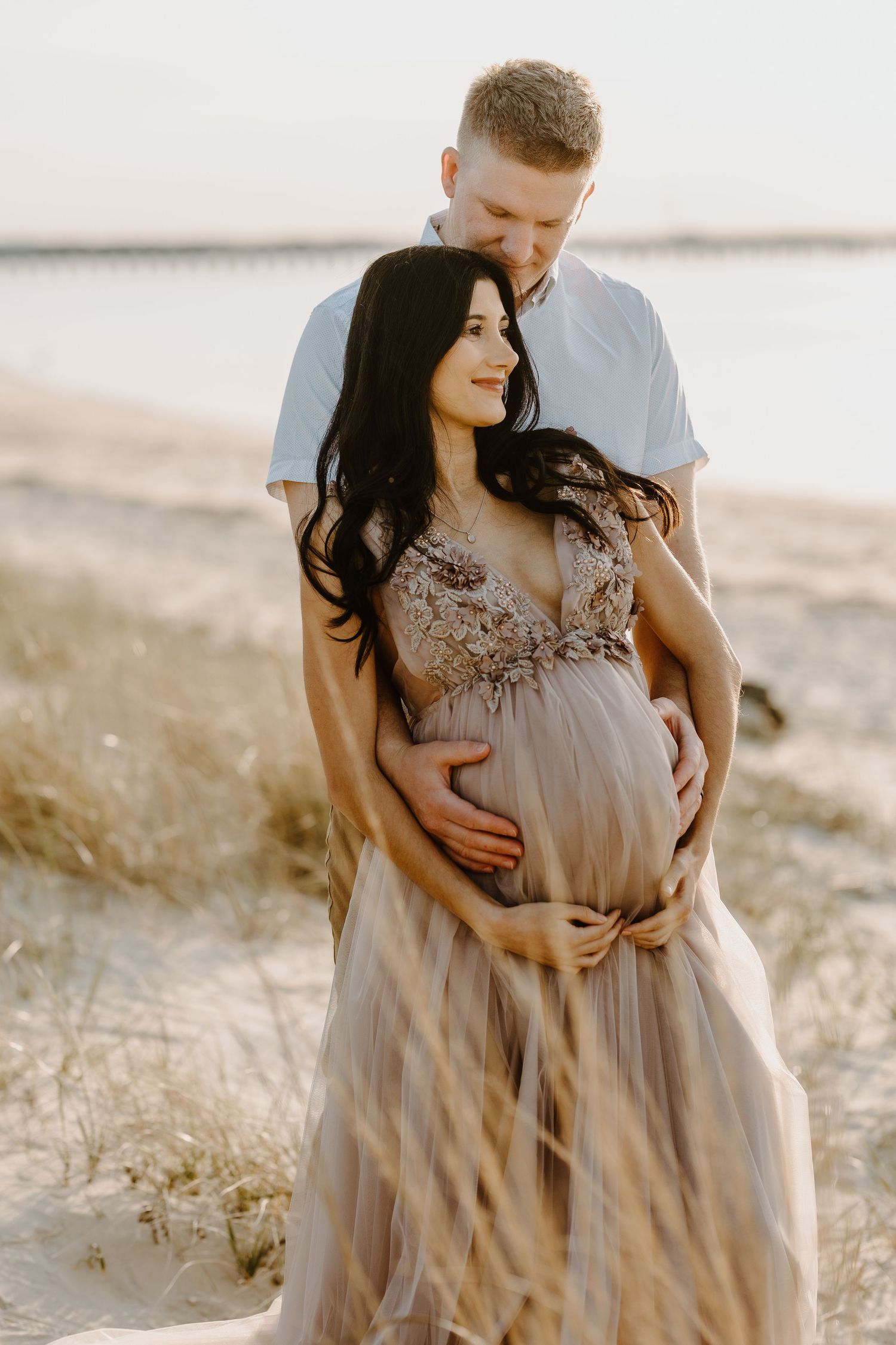 Pregnant couple embracing on a sandy beach at sunset, woman in elegant floral tulle maternity gown.