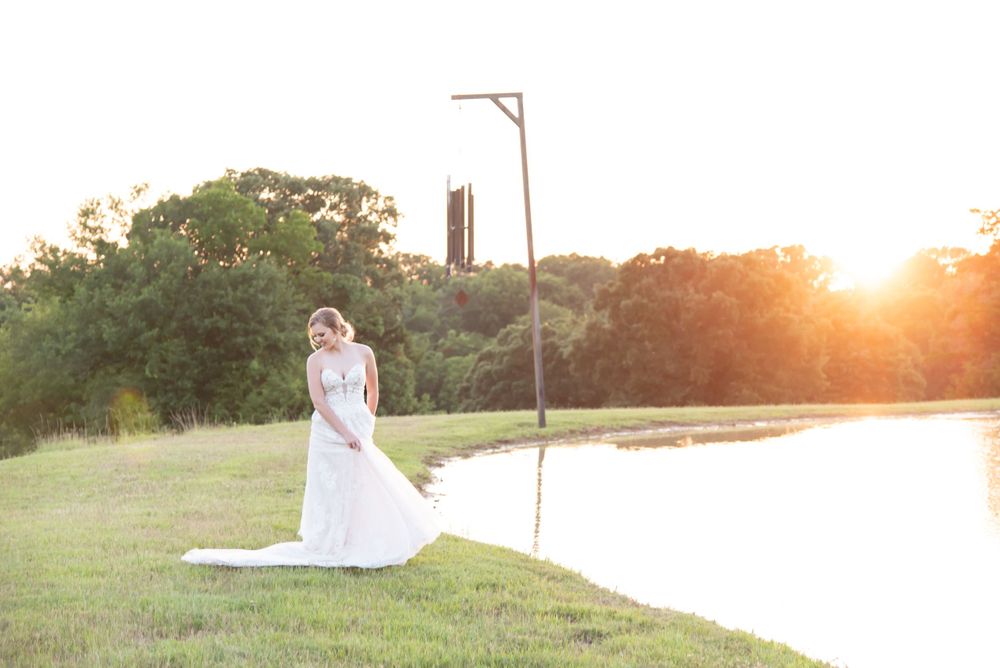 A bride in white gown poses by a lake during golden hour sunset.