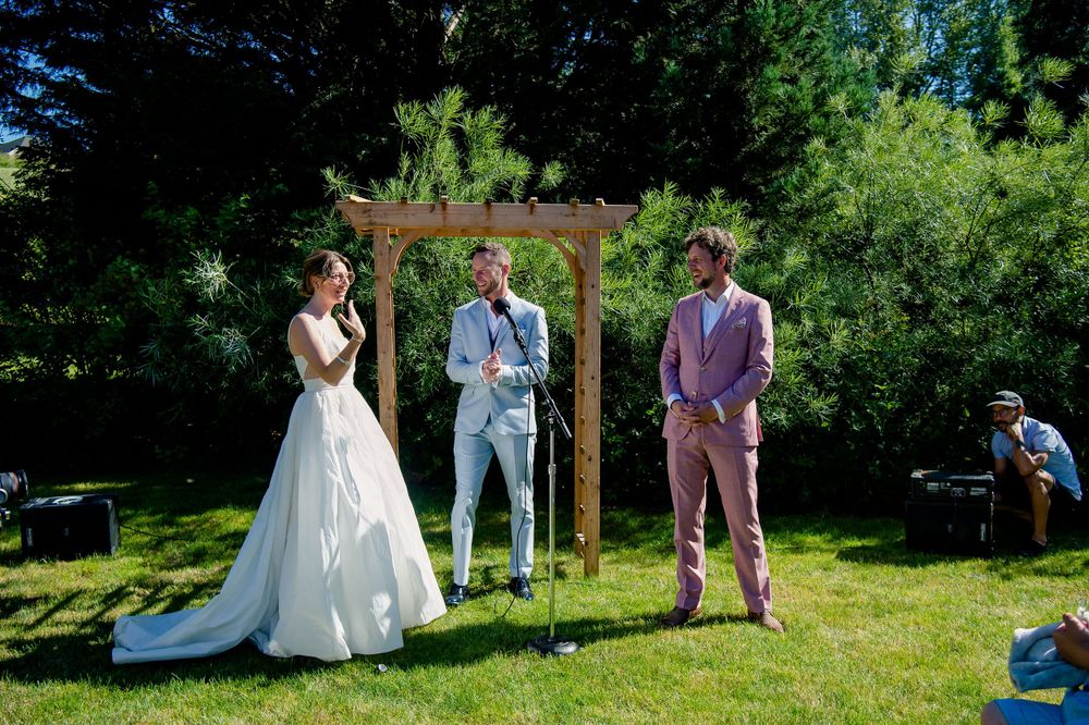 Wedding ceremony under a wooden arch with three people in formal attire on lush green lawn.