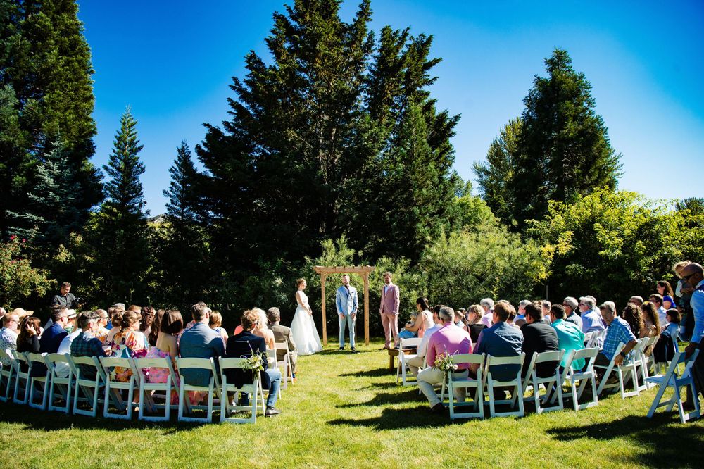 Outdoor wedding ceremony setup with white chairs on grass surrounded by evergreen trees under blue sky.