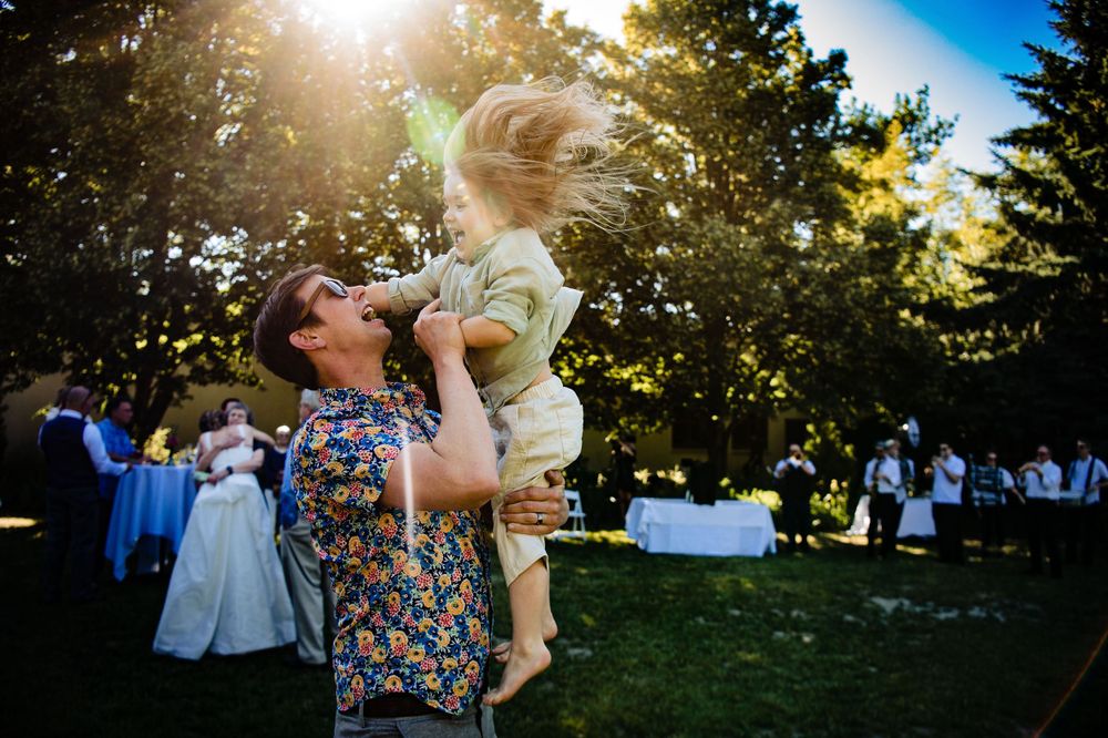 A tender backlit moment at an outdoor reception as guests celebrate in the background.