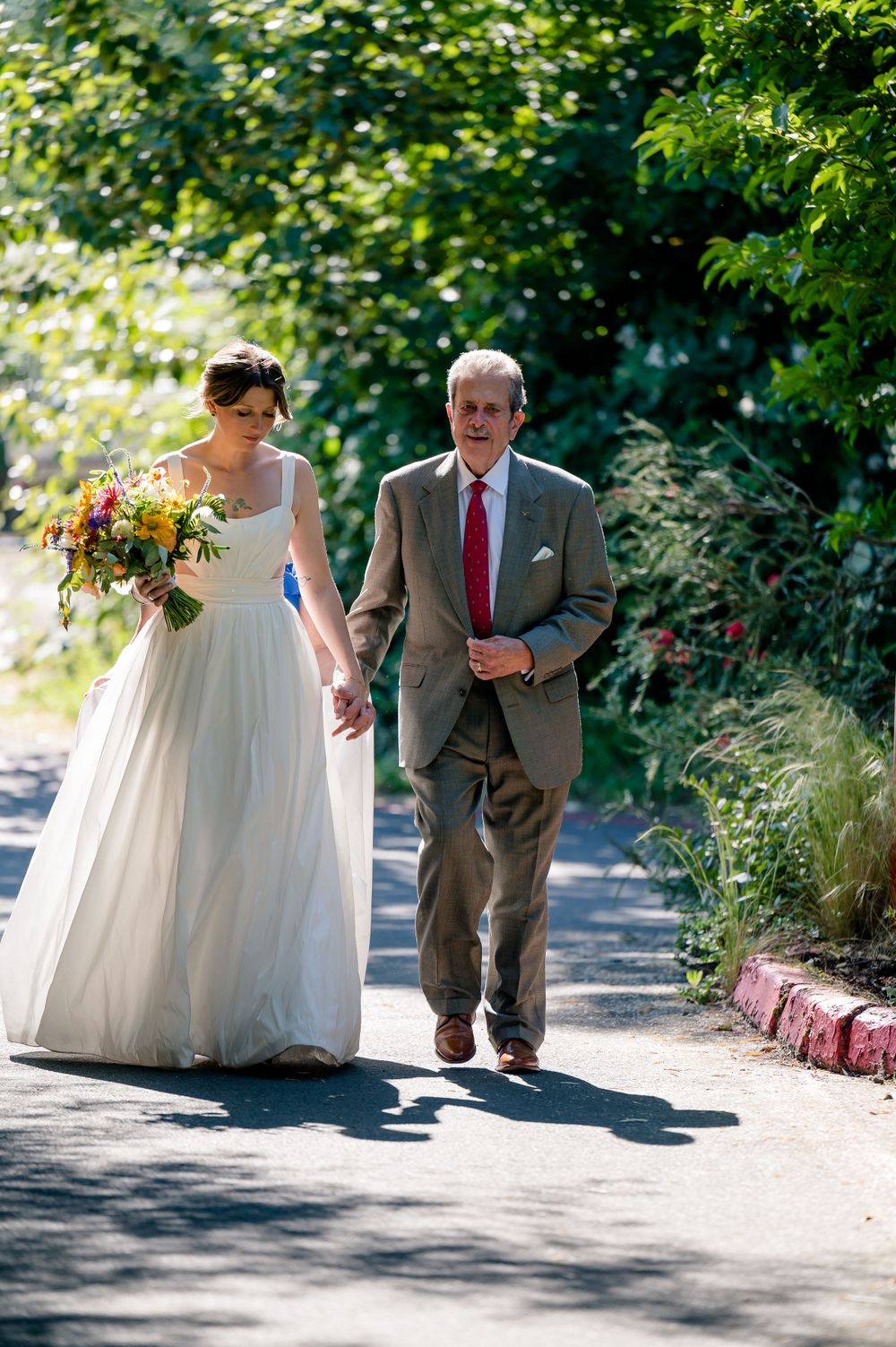 Wedding day walk along a sunny garden path with colorful bouquet and formal attire.