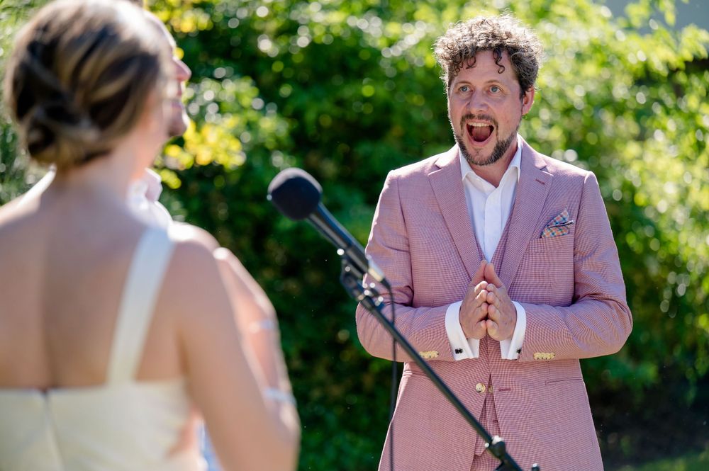 A couple exchanges vows during an outdoor summer wedding ceremony with the groom wearing a pink suit.