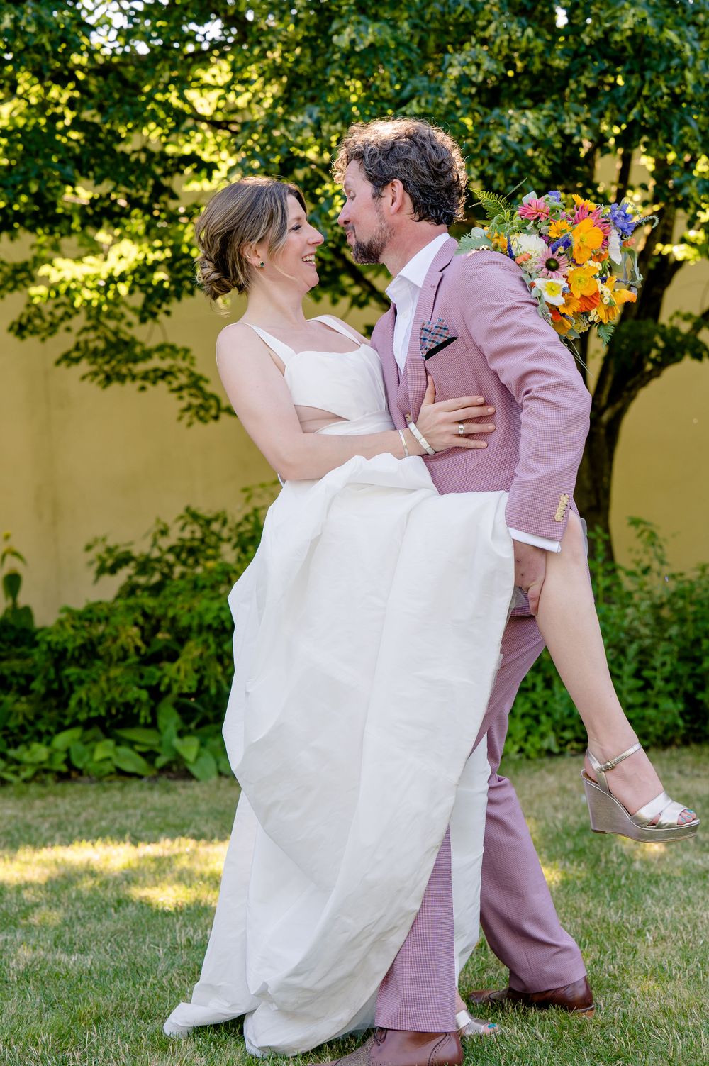 Wedding couple in white dress and pink suit share intimate dance moment in garden setting.