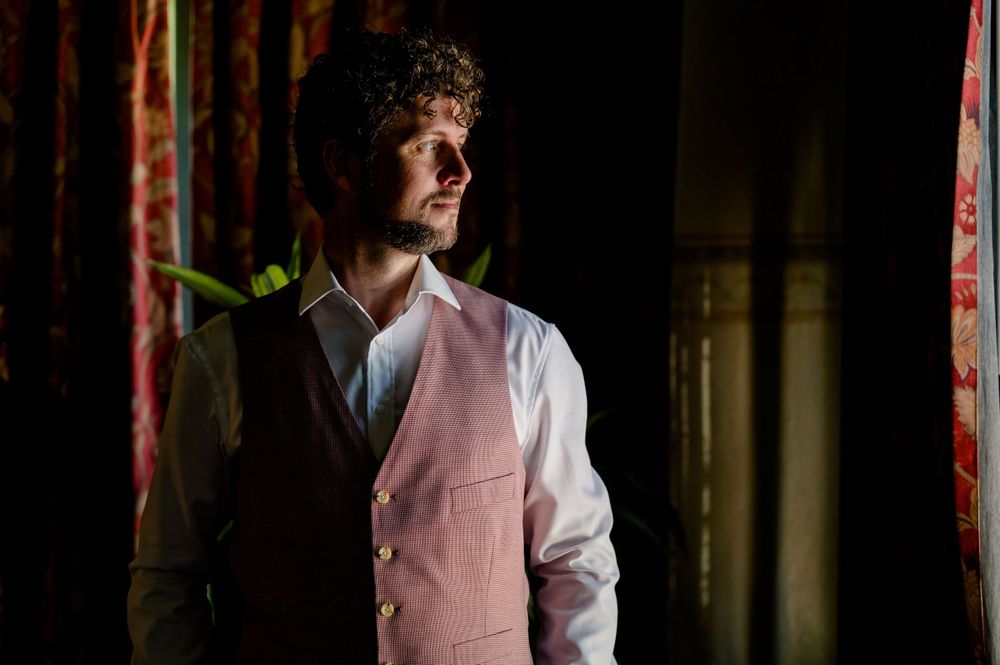 A groom in a pink vest and white shirt stands in dramatic lighting against a dark background.