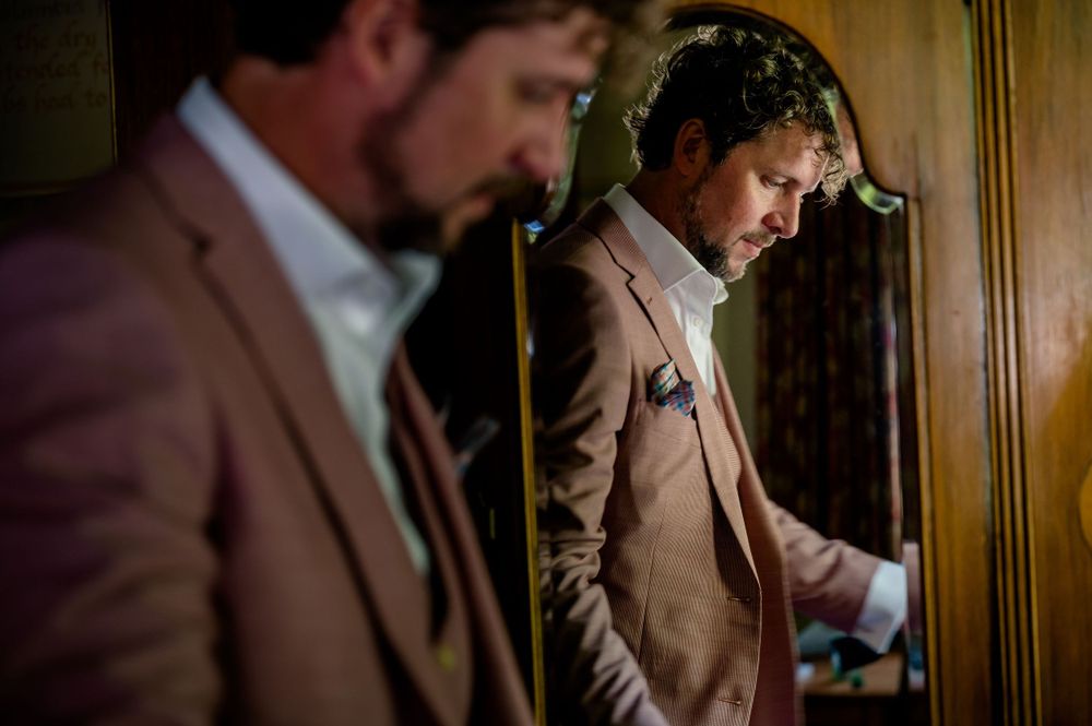 A man in brown suit adjusts his tie while looking in mirror.