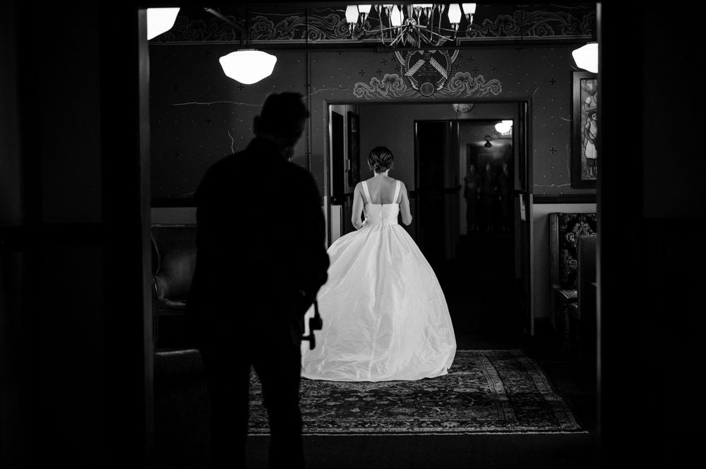A silhouetted figure watches a bride in white gown through doorway.