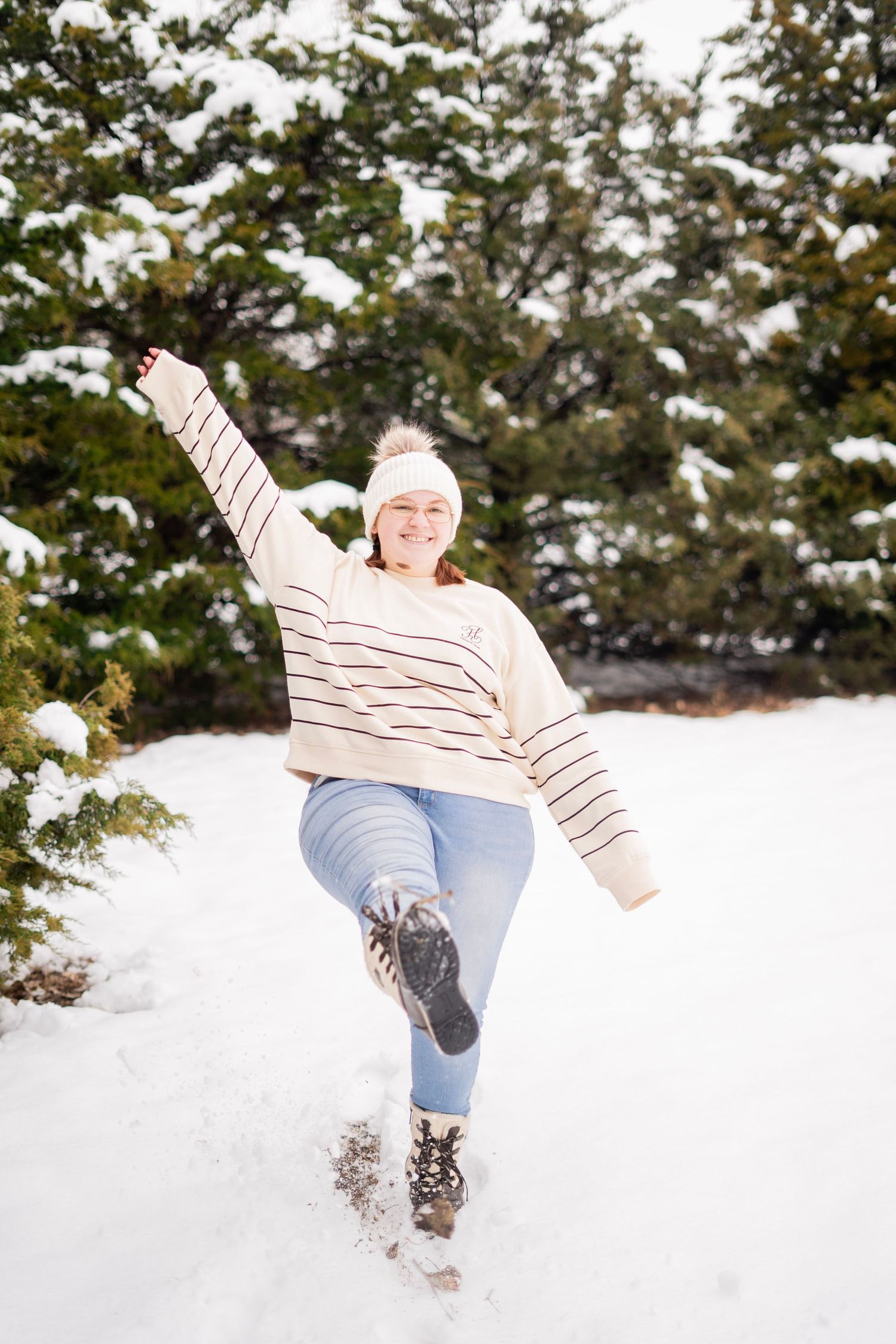 Cheerful woman in striped sweater and jeans kicks leg up playfully in snowy evergreen forest.