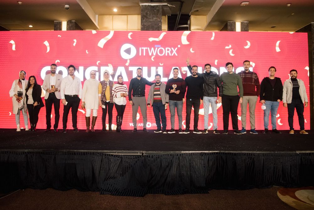 Group photo of team members on stage with red backdrop and congratulatory message.