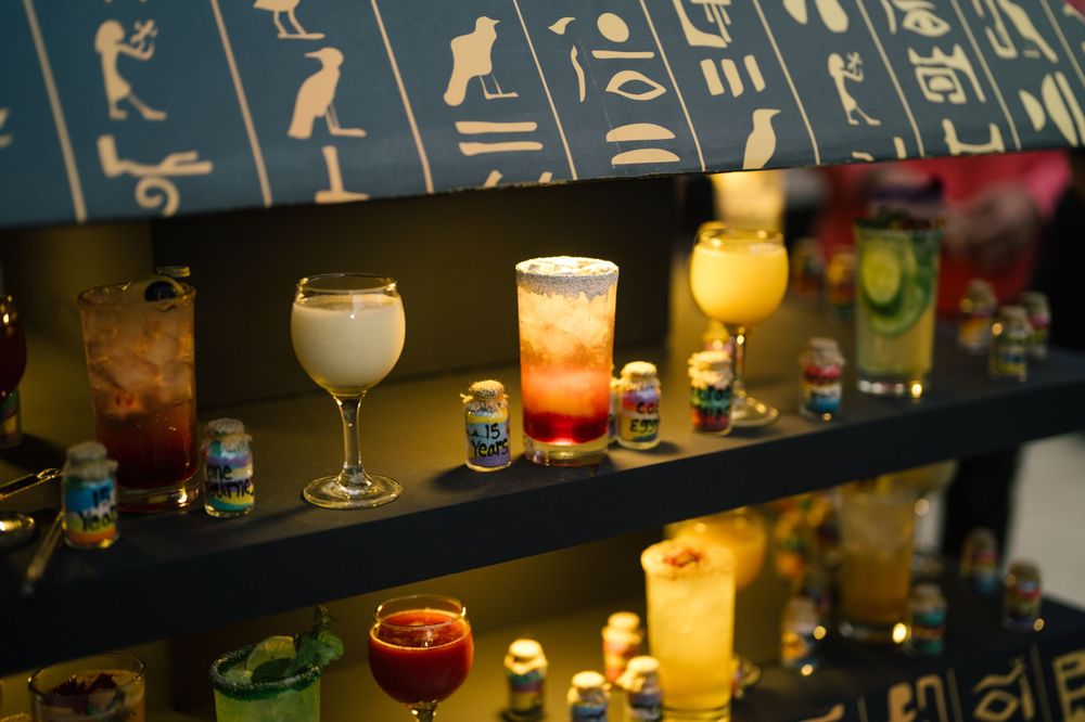 Colorful cocktails and beverages displayed on a dark shelf with Egyptian hieroglyphic decorations above.