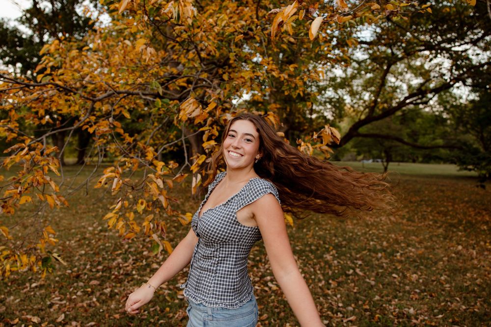 A woman spins joyfully in a park surrounded by autumn leaves and golden sunlight.