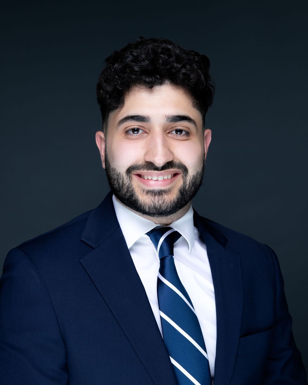 Professional headshot of a smiling person wearing a navy suit and striped tie against a dark background.