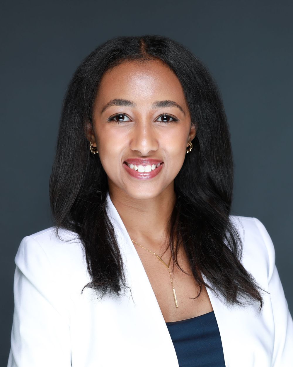 Corporate portrait with person wearing white blazer and navy top against grey backdrop.
