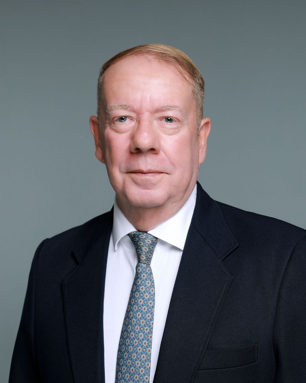 Professional headshot of a senior businessman in a dark suit and patterned tie against a gray background.