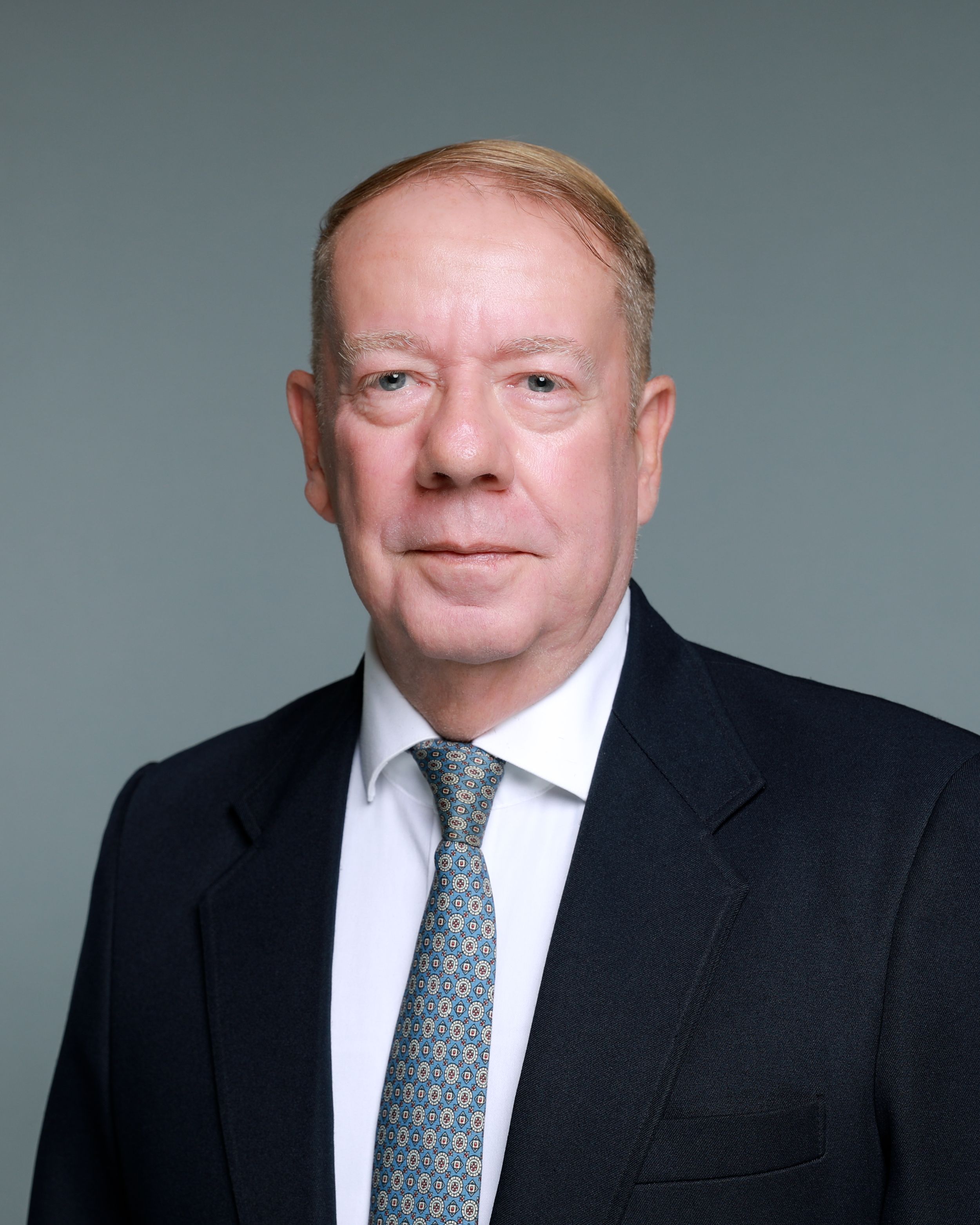 Professional headshot of a senior businessman in a dark suit and patterned tie against a gray background.
