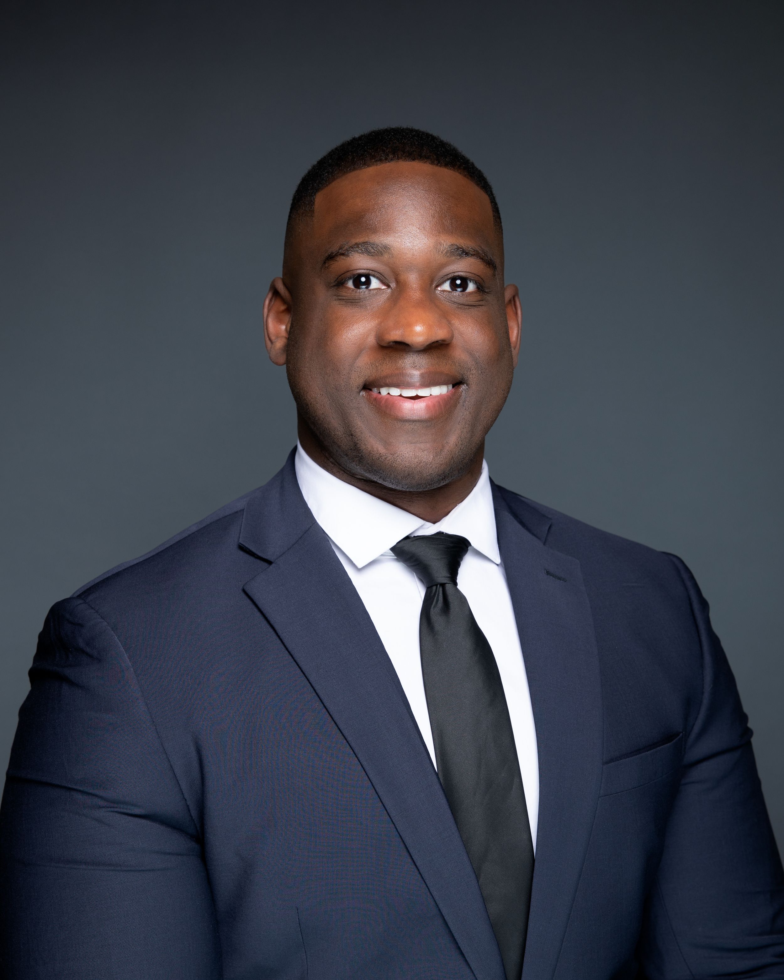 Professional headshot of a smiling man in a navy blue suit and black tie against a dark gray background.