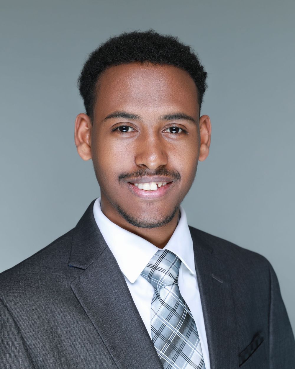 Professional headshot of a young professional in a dark suit and blue plaid tie against a gray background.