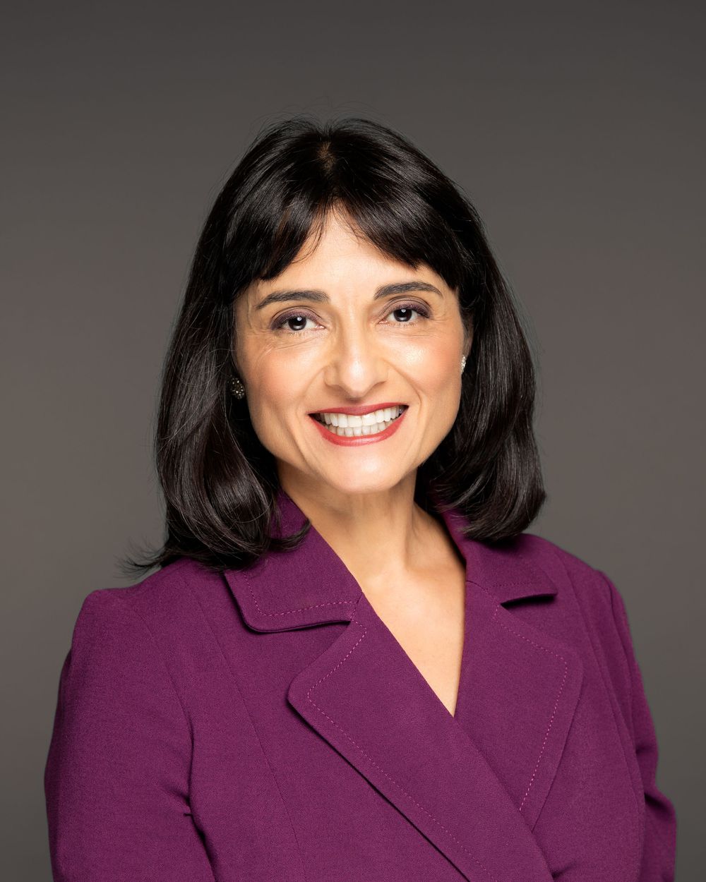 Professional headshot of a smiling woman with dark hair wearing a purple blazer against a gray background.