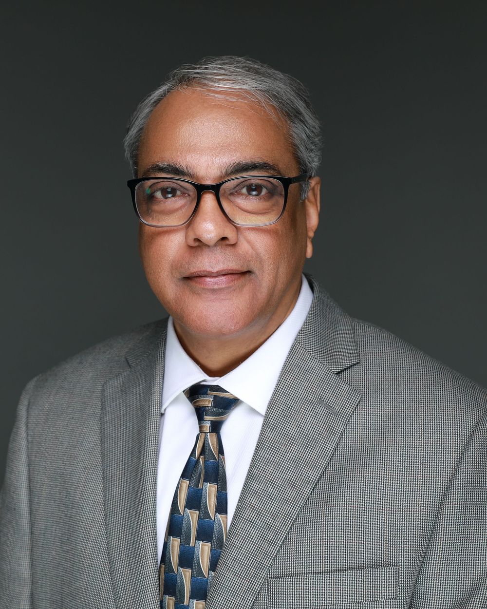 Professional headshot of a person wearing glasses, a gray suit, and patterned tie against a dark background.