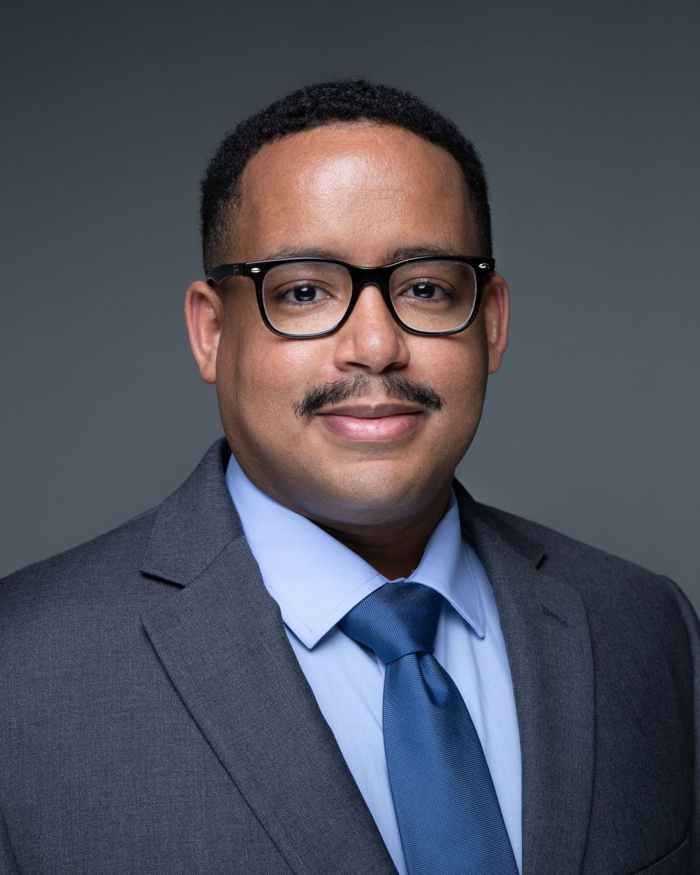Professional headshot of a man in a gray suit and blue tie wearing glasses with a warm smile.