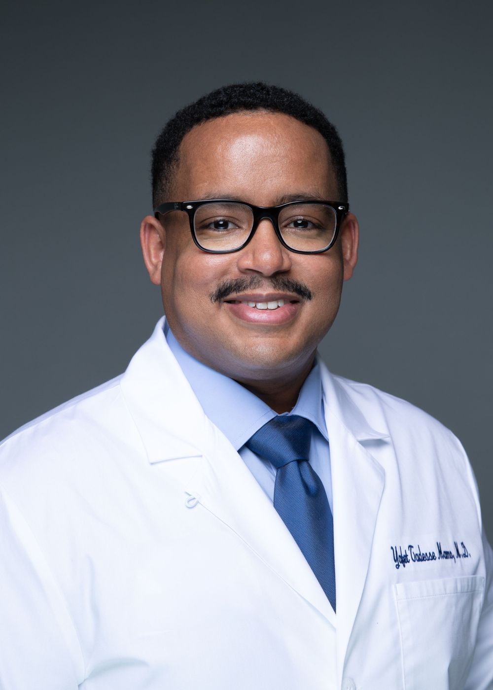 Professional portrait of a smiling doctor in a white coat, blue shirt, and tie wearing black-framed glasses.