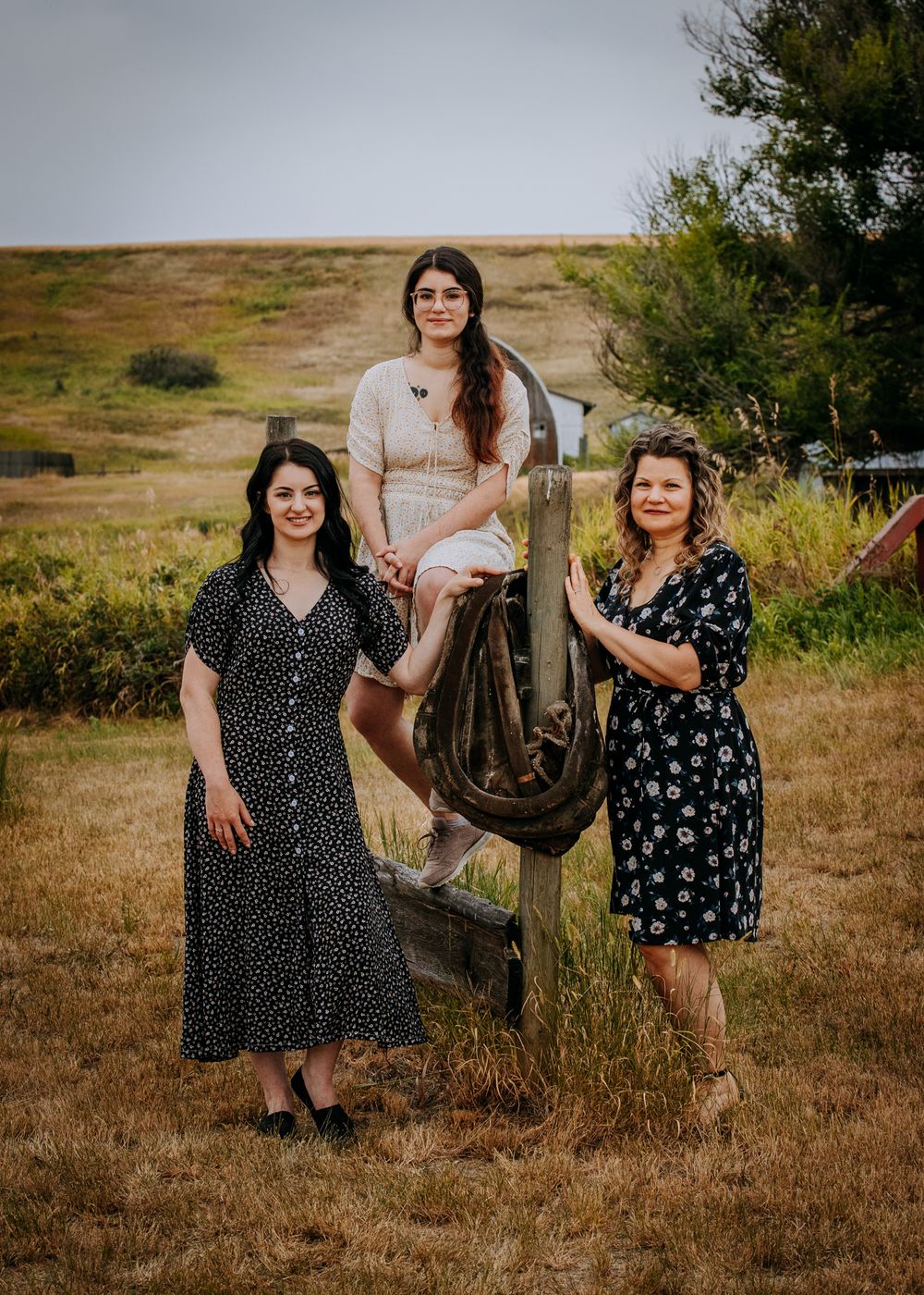 Three friends in floral dresses pose together near an old wooden fence post in a rural setting.