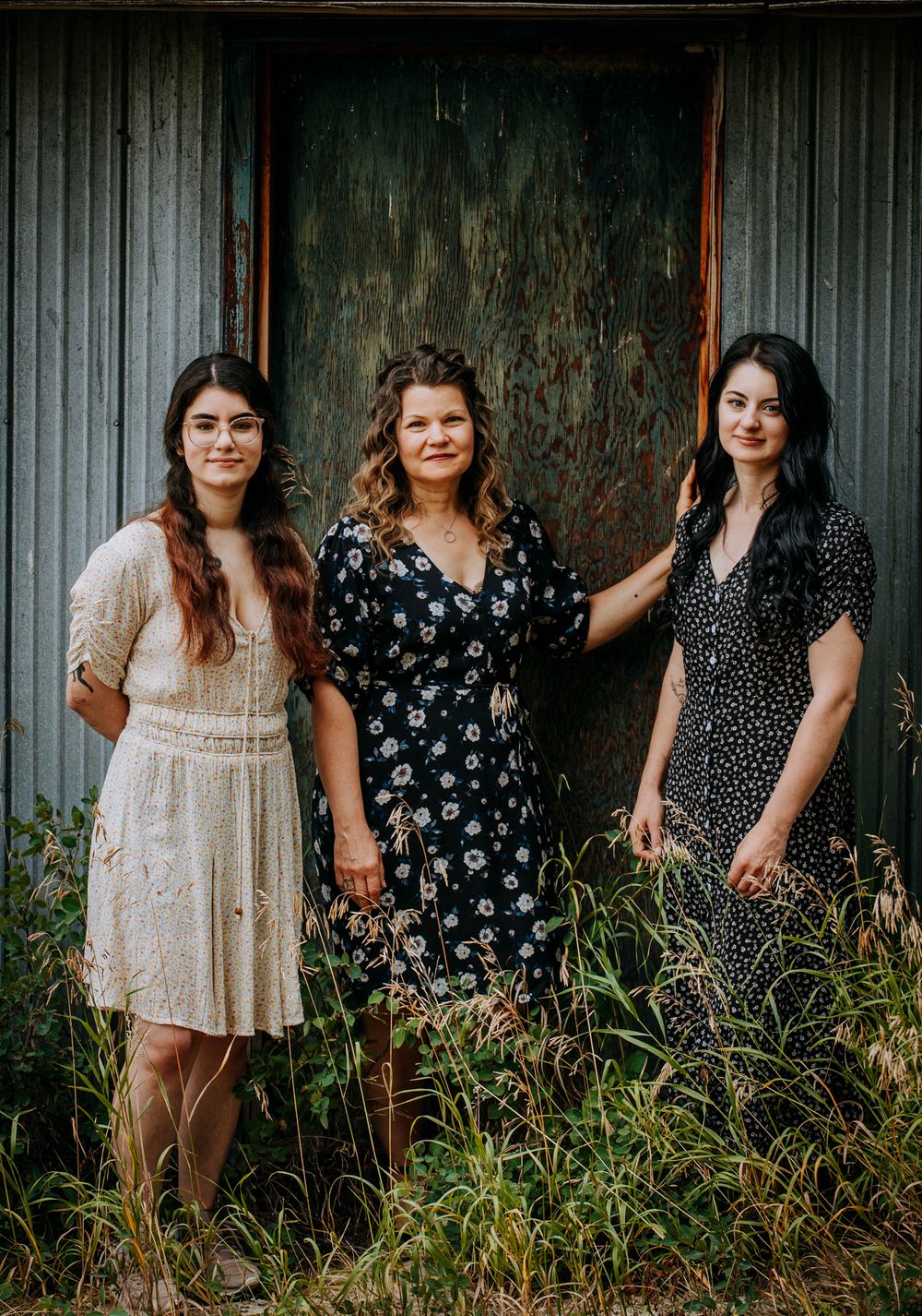 Three friends in vintage-style dresses pose together against a rustic metal wall backdrop.