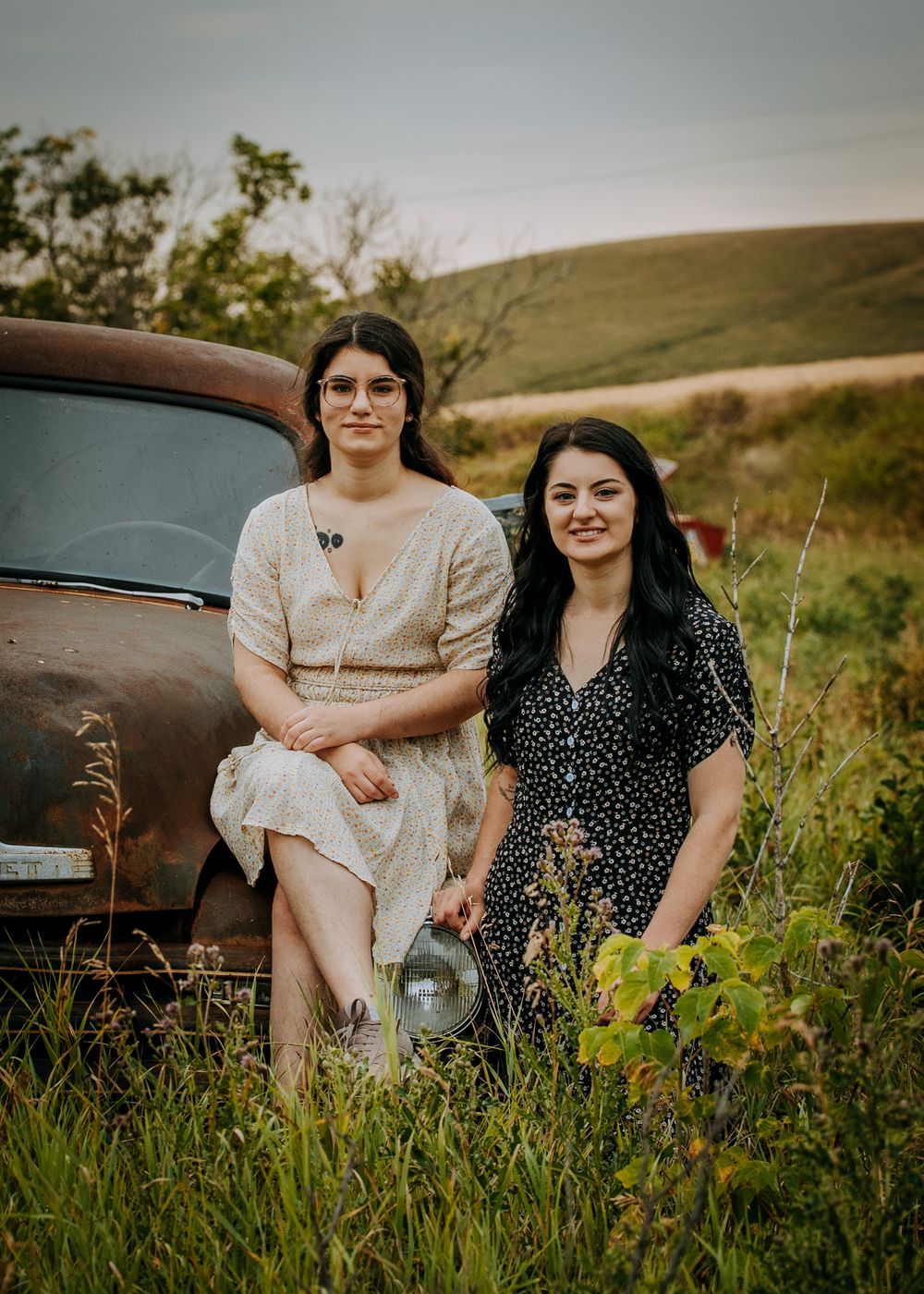 Two friends in contrasting dresses sit by a vintage rusty truck in a grassy field.