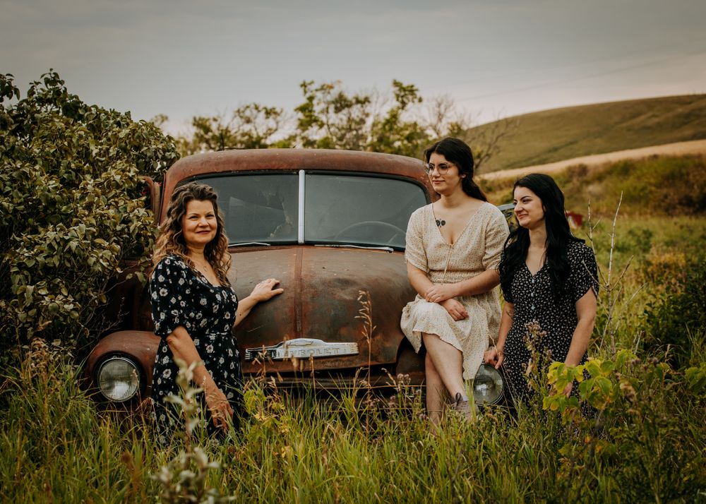 Friends in vintage dresses pose together with an old rusty truck in a rural field setting.