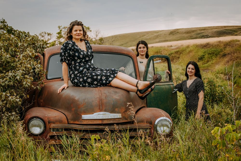 Three people pose playfully with an antique rustic truck in a rolling countryside setting.