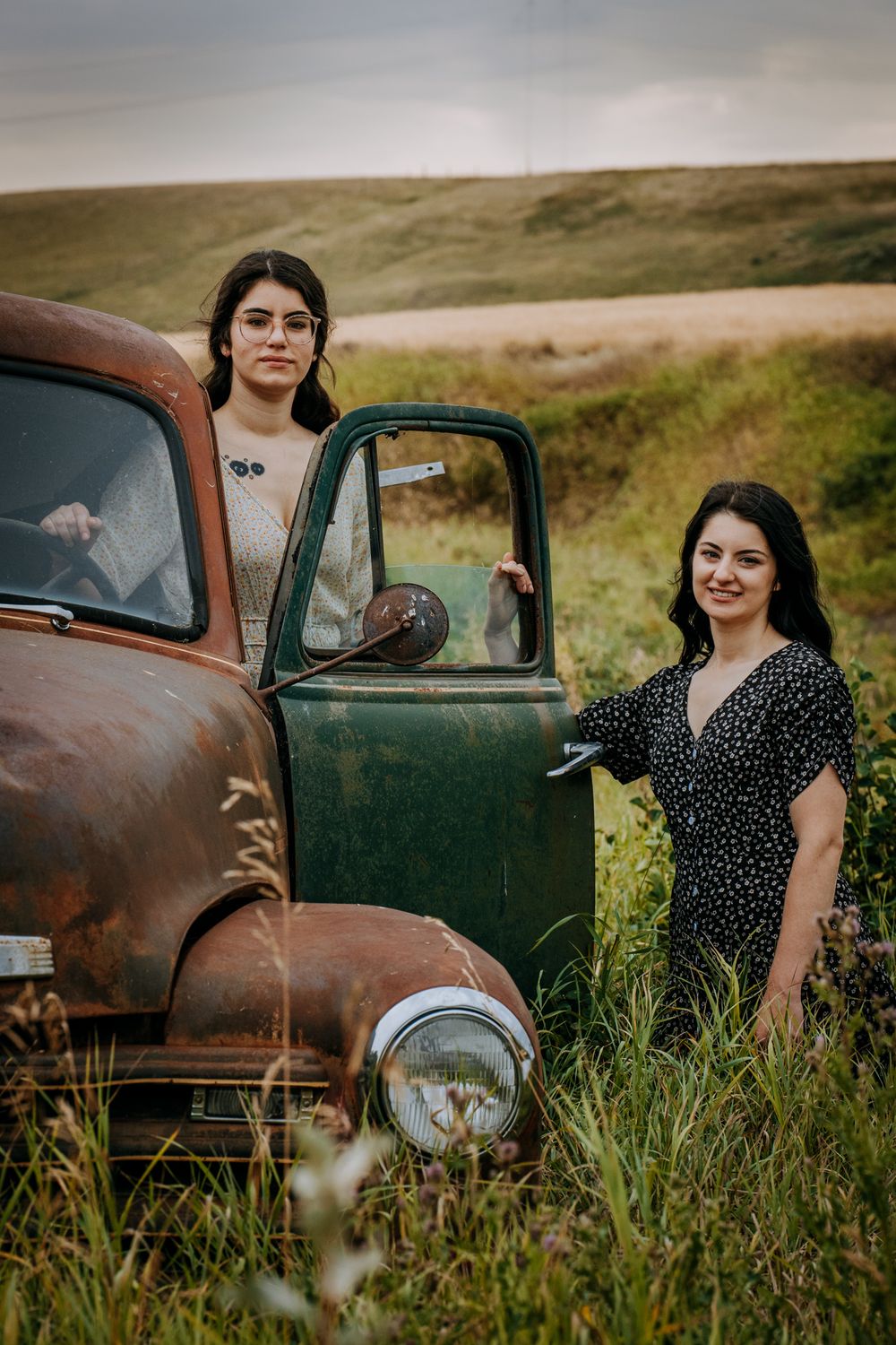 Two people pose beside a rustic vintage truck in a grassy field at sunset.