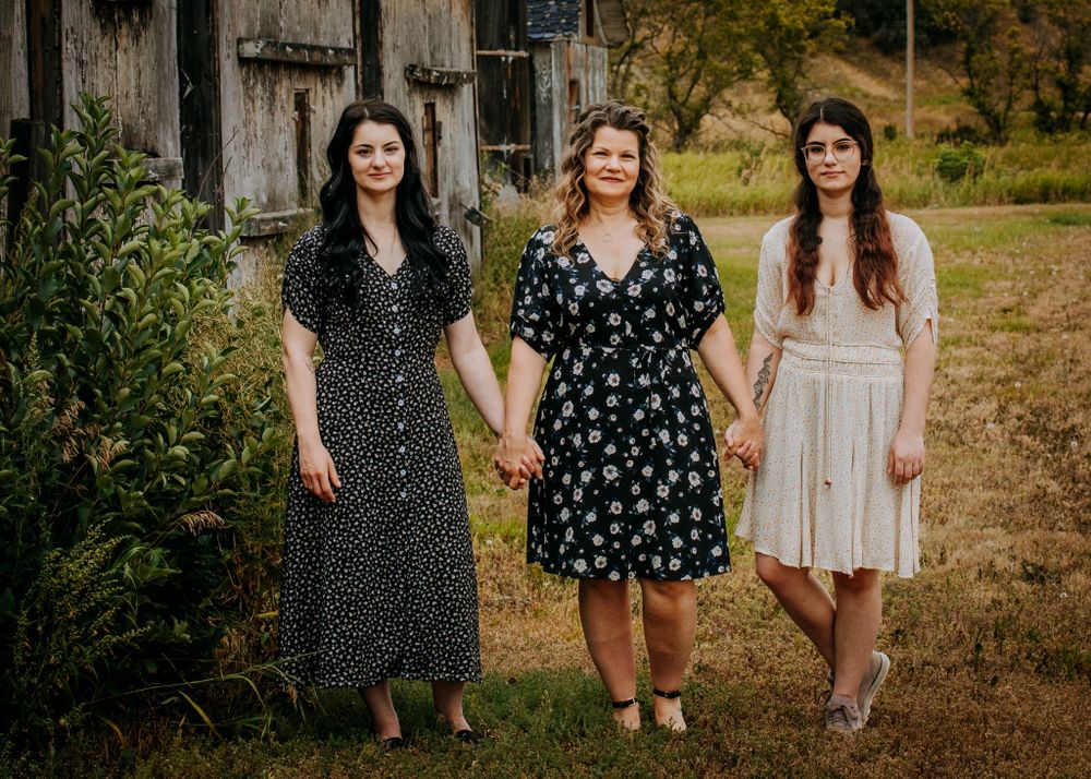 Three friends in patterned dresses hold hands while standing together near a rustic barn.