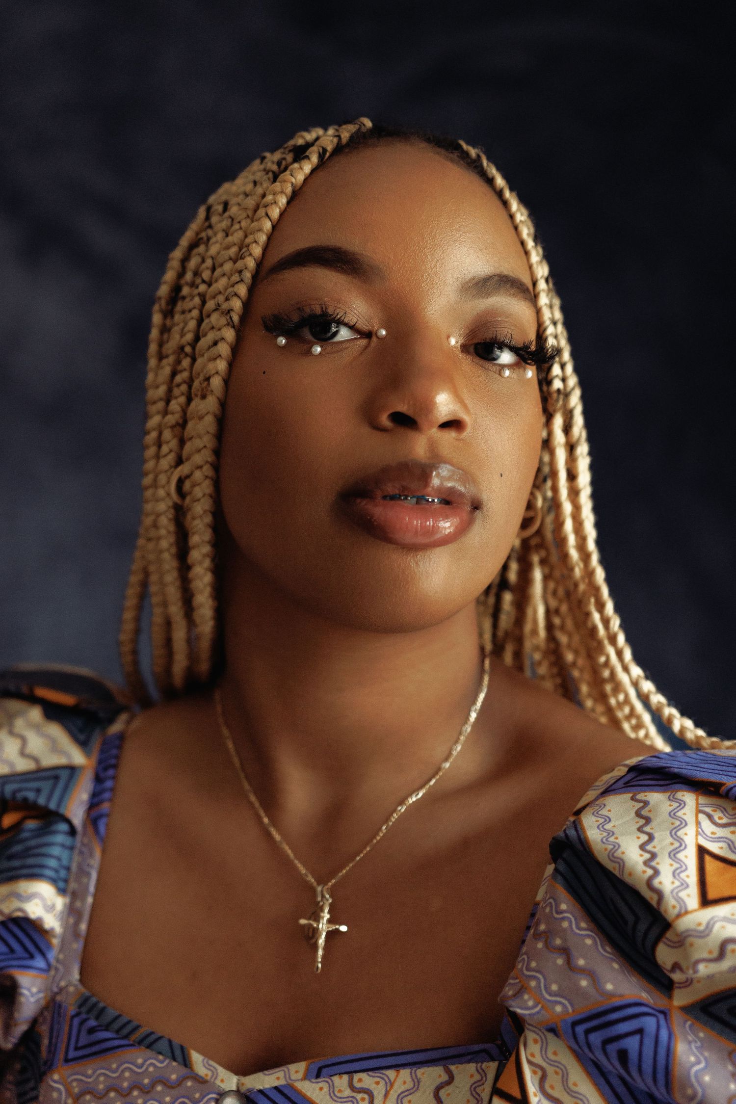 Portrait with blonde braids and cross necklace against dark background.