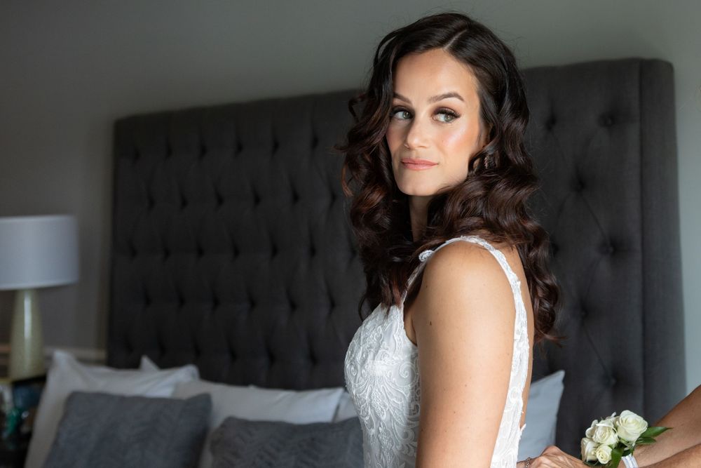 Beautiful bride in white lace dress with elegant curled hair posing in bedroom.