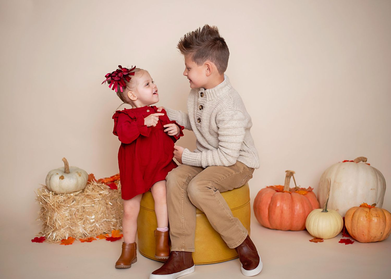 Two children pose with pumpkins and hay bales in a fall-themed studio setting.
