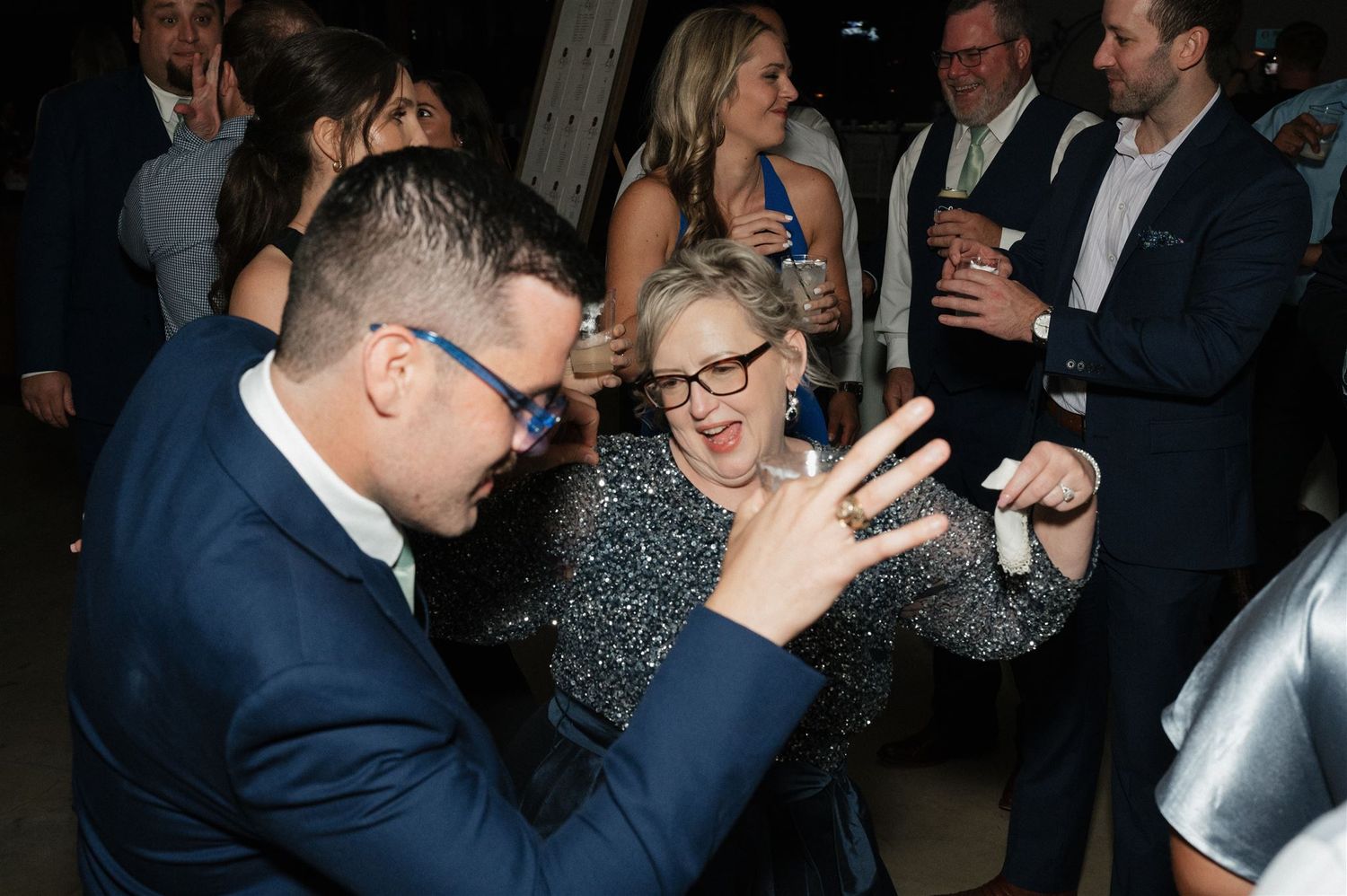 Wedding guests dancing and celebrating in formal attire at a dimly lit reception.
