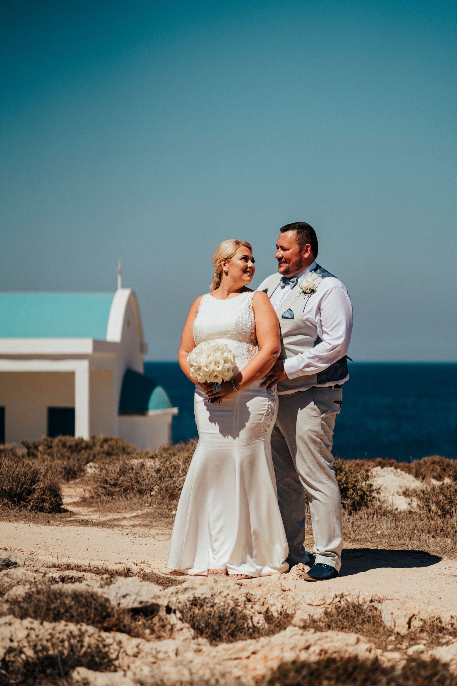 A wedding couple shares a joyful moment against a stunning coastal backdrop with a white chapel.