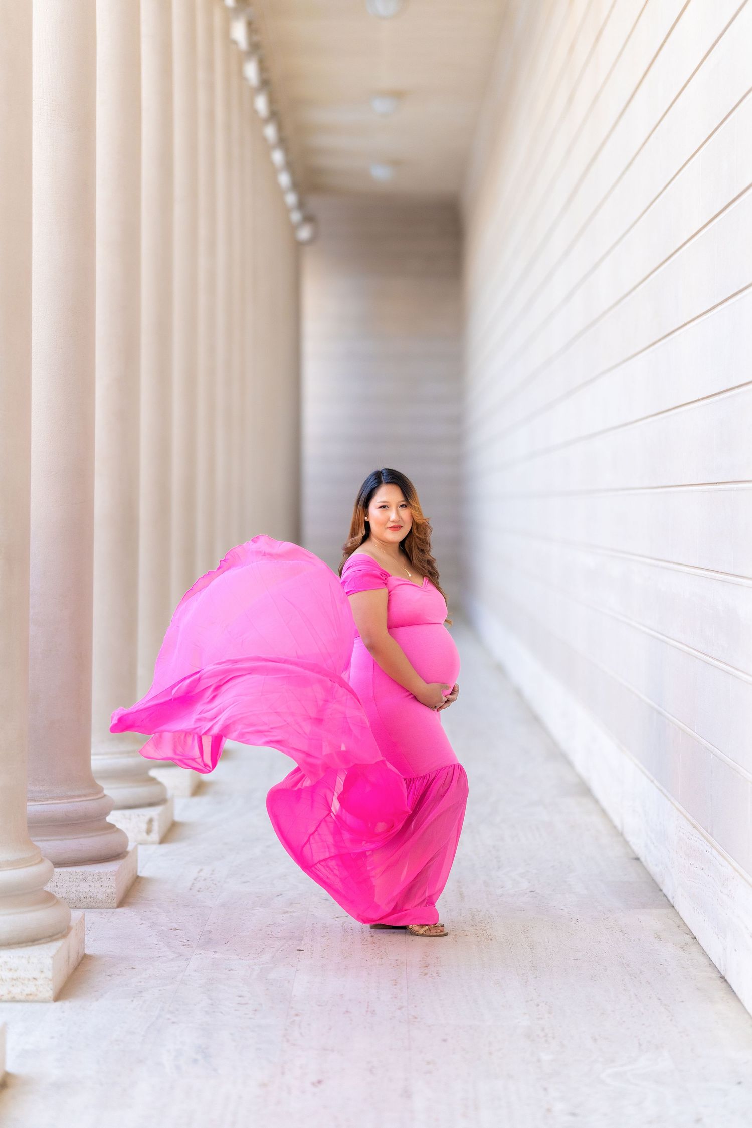 A maternity photo of a flowing pink dress in a hallway with classical columns.