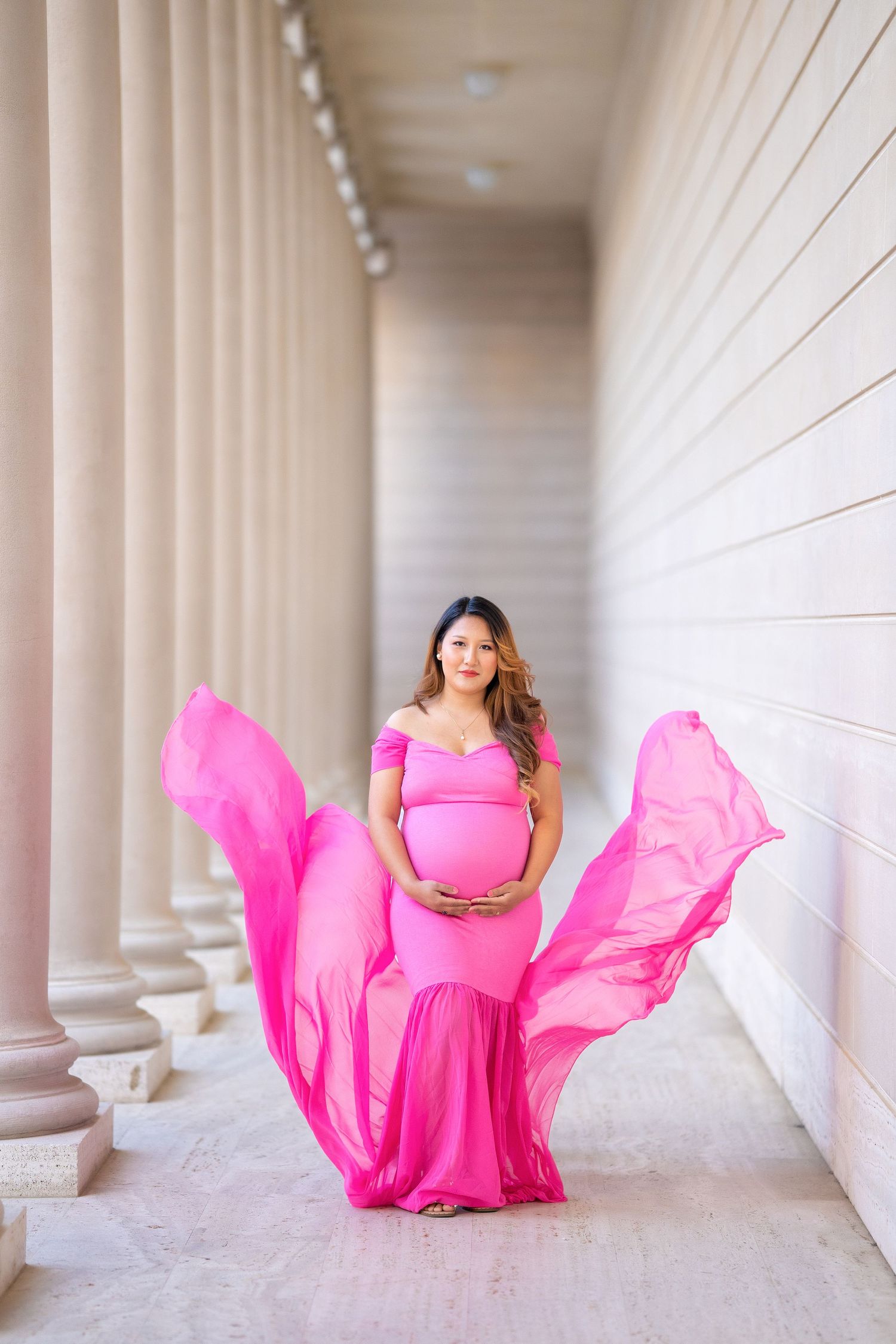 A maternity portrait with a flowing pink dress in an elegant columned corridor.