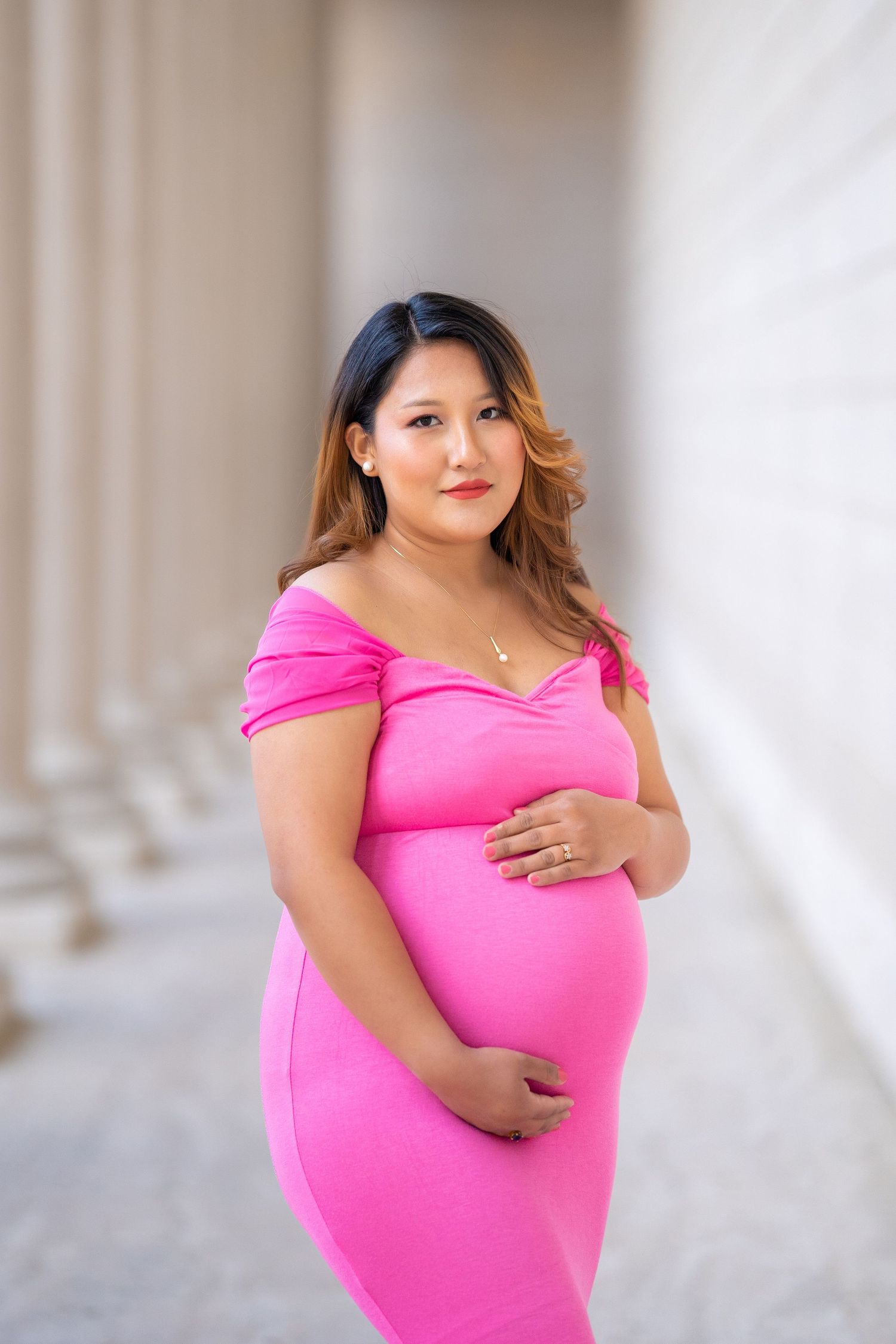 A maternity portrait featuring a pink form-fitting dress in a classic architectural setting.