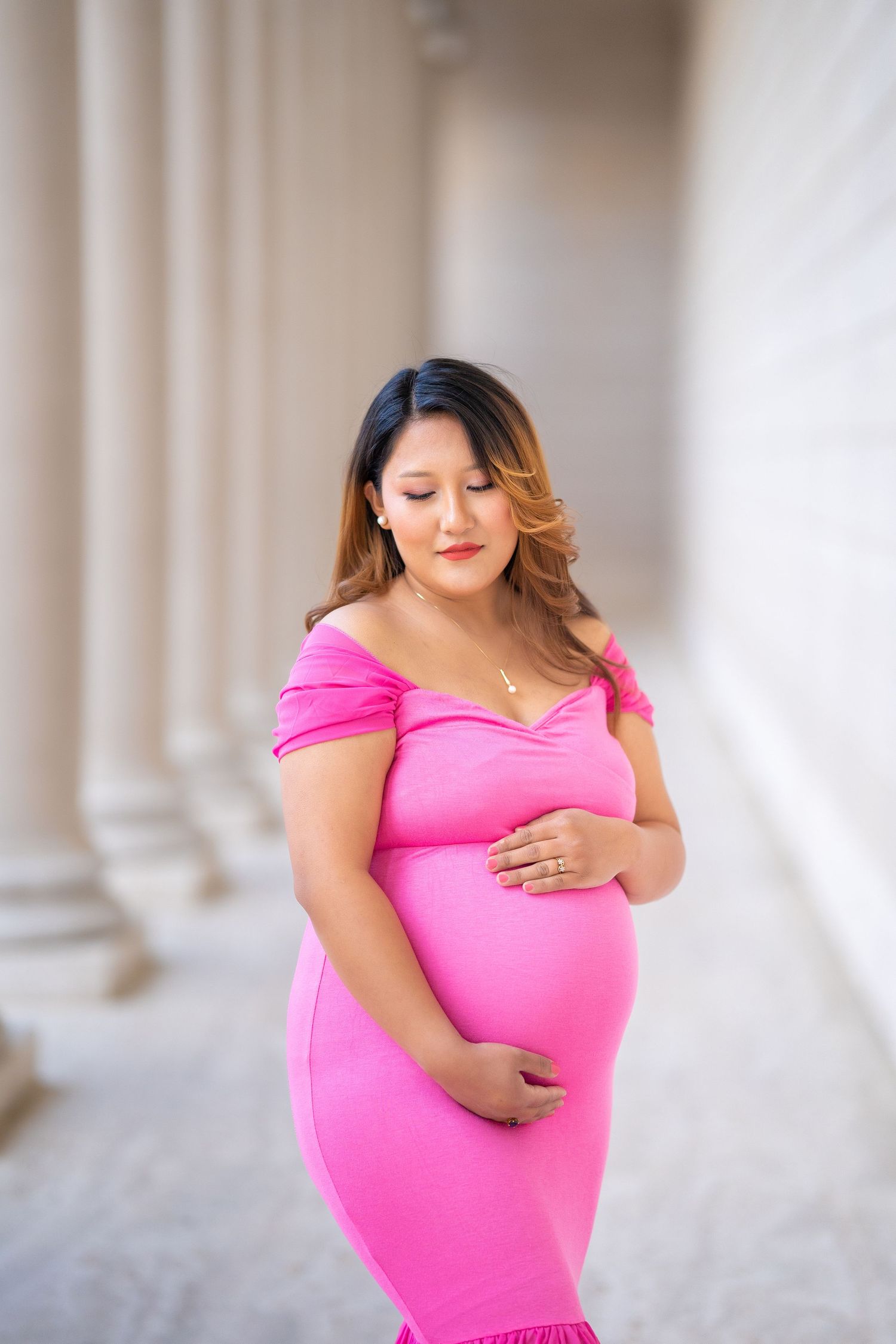 A maternity portrait against white walls and columns in a fitted pink dress.