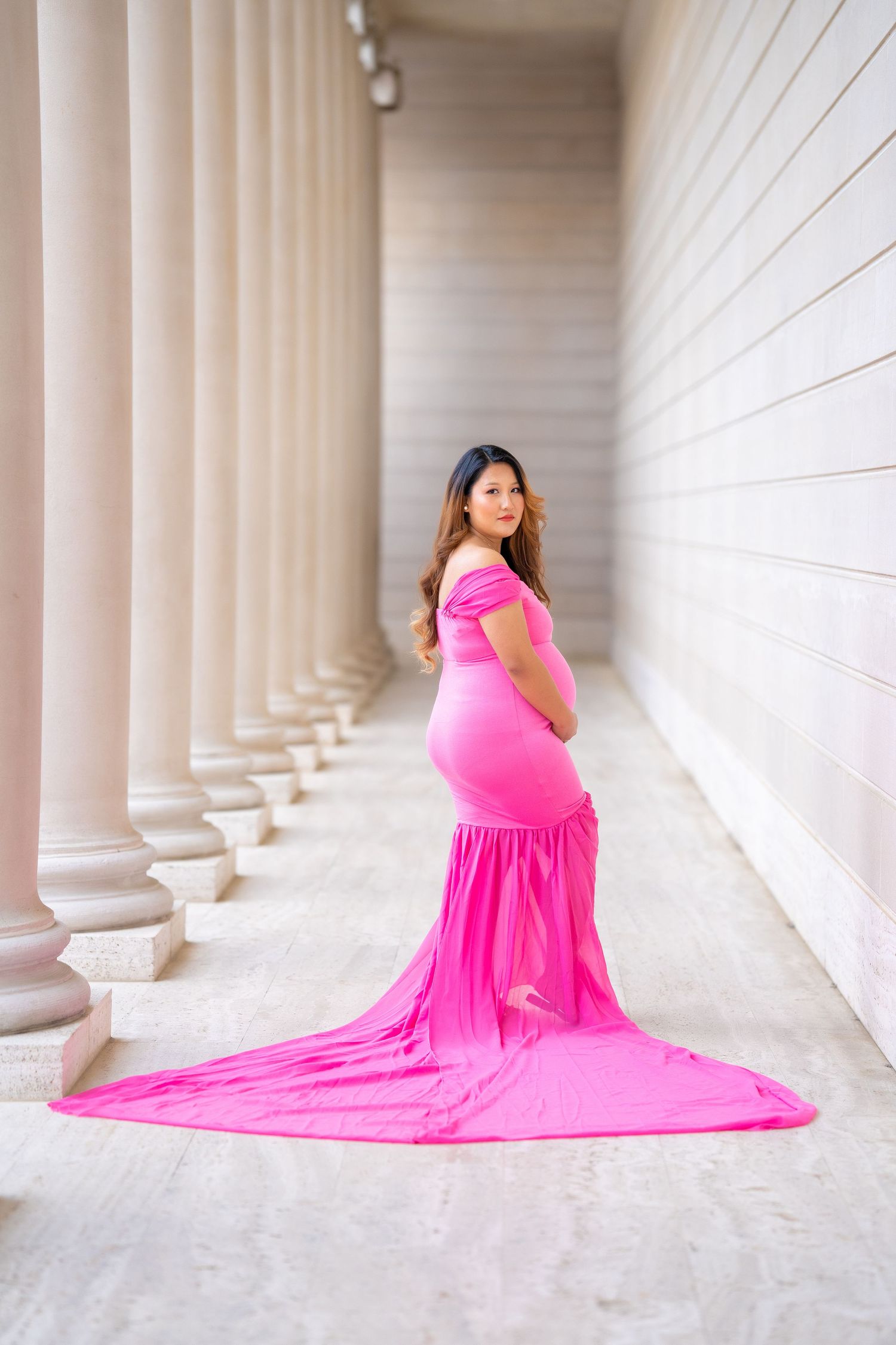 A dramatic maternity photo with a trailing pink dress against classical architecture.