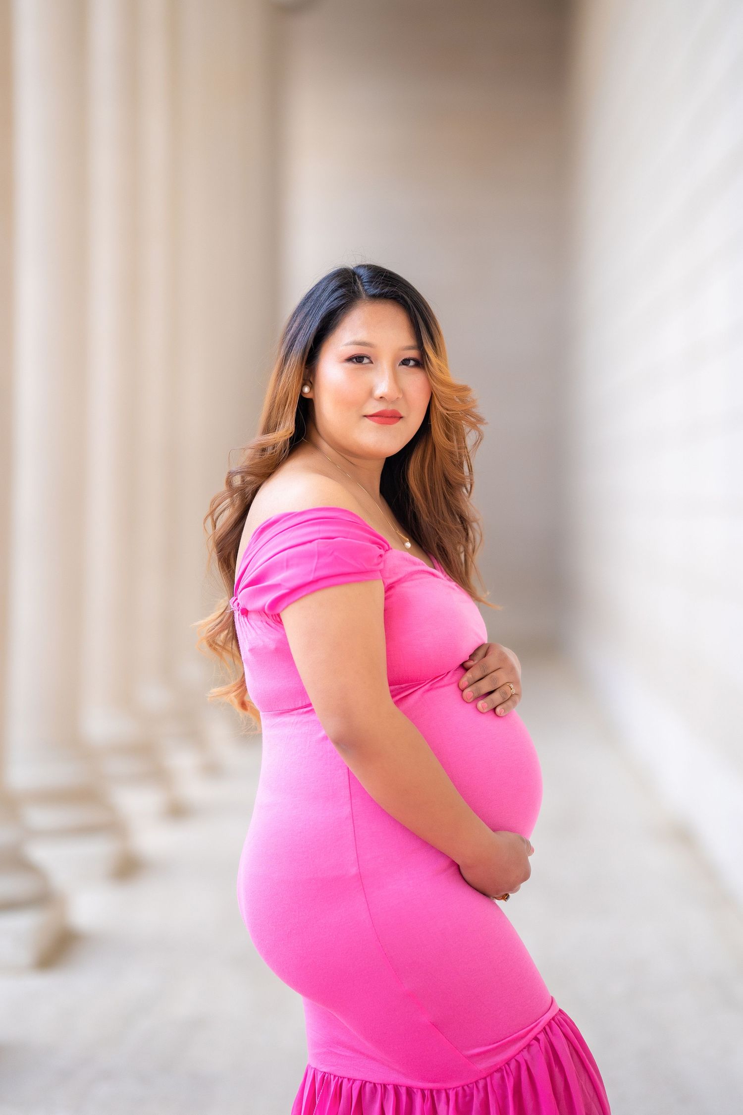 A side-view maternity portrait in a fitted pink dress against classical columns.